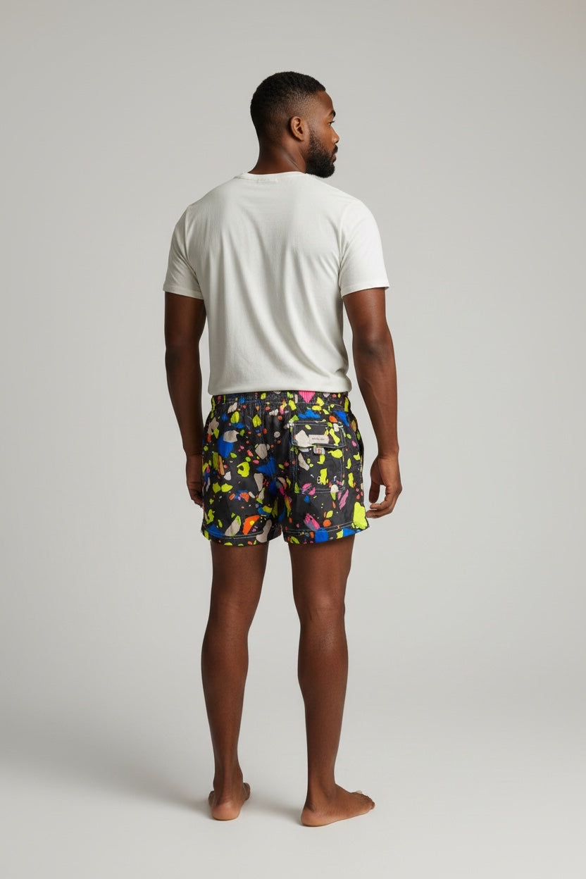 98 Coast men's black swim shorts with vibrant multicolor paint splatter print, elastic waistband and drawstring, back pocket detail, model, white t-shirt, back view