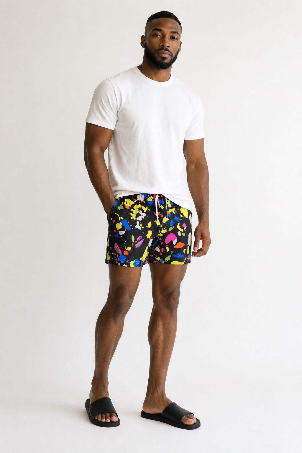 98 Coast men's black swim shorts with vibrant multicolor paint splatter print, elastic waistband and drawstring, back pocket detail, model, white t-shirt, front view